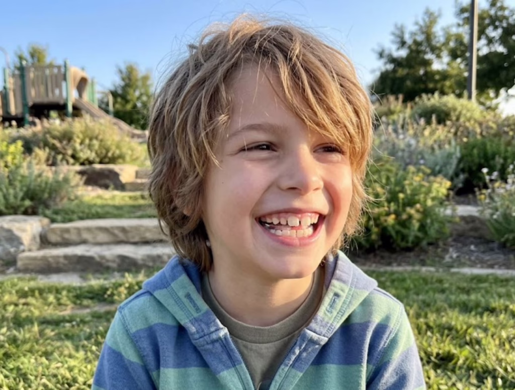 A happy young boy with a wide, healthy smile sitting outdoors in Mansfield, TX, representing the successful results of airway-focused palate expansion treatment at Central Park Dental & Orthodontics.